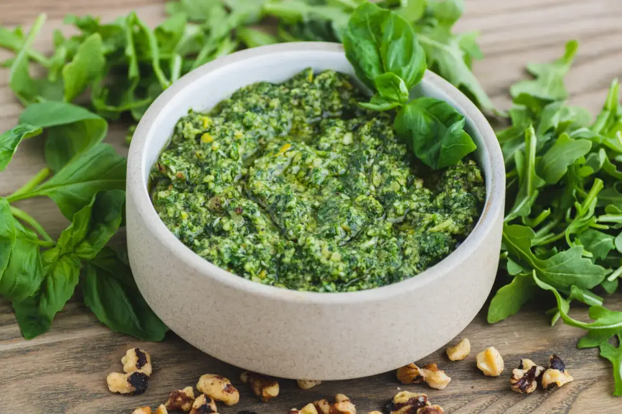 Schüssel mit frischem Rucola Pesto, umgeben von Rucola-Blättern und Walnüssen auf einem Holztisch.