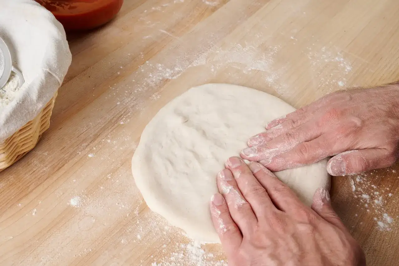 Hände formen pizzateig auf bemehltem Holztisch. Zutaten für Pizza sind bereit.