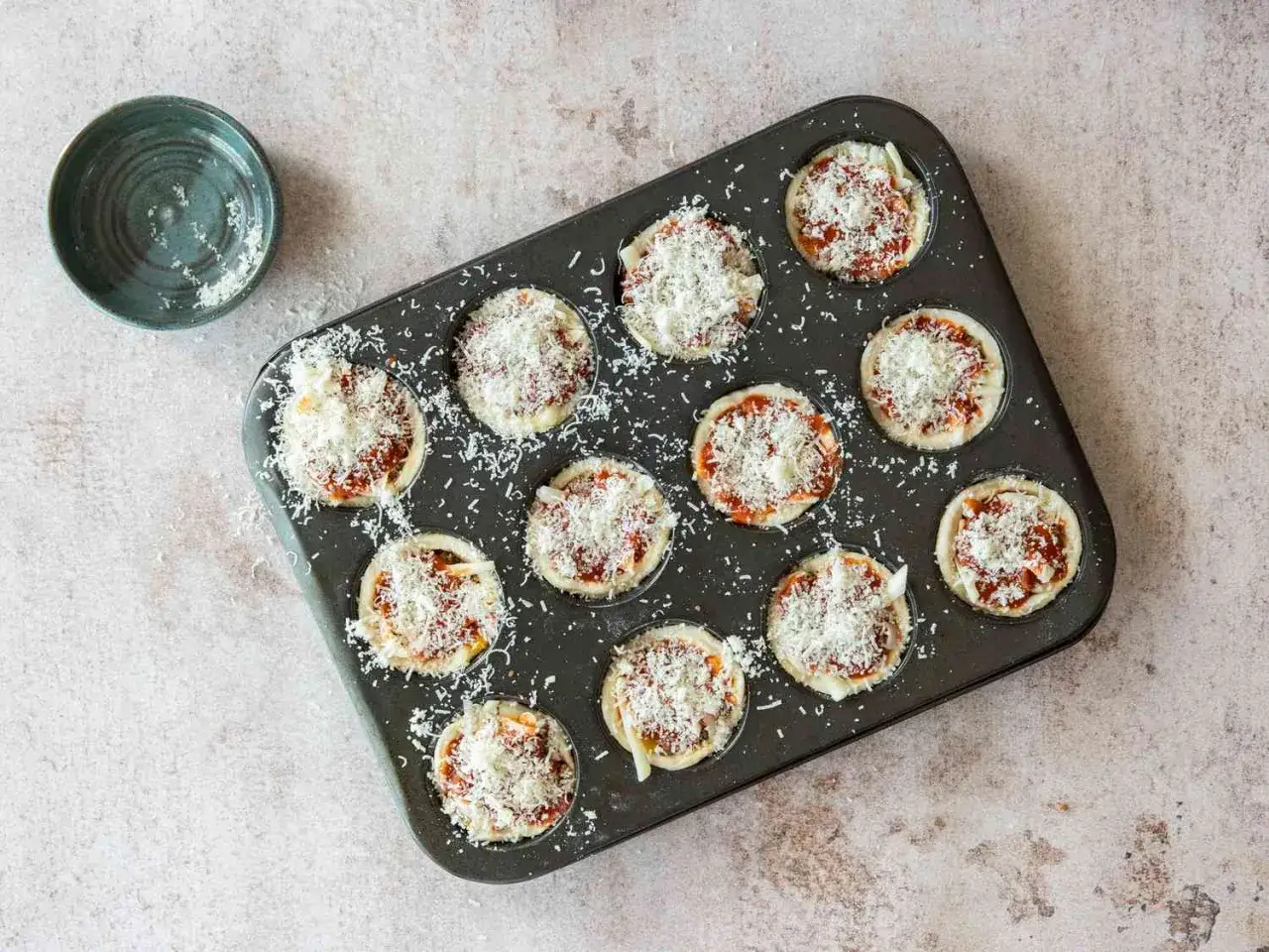 Zw&ouml;lf kleine Pizza Muffins in einer Muffinform, bereit zum Backen, mit geriebenem K&auml;se bestreut.