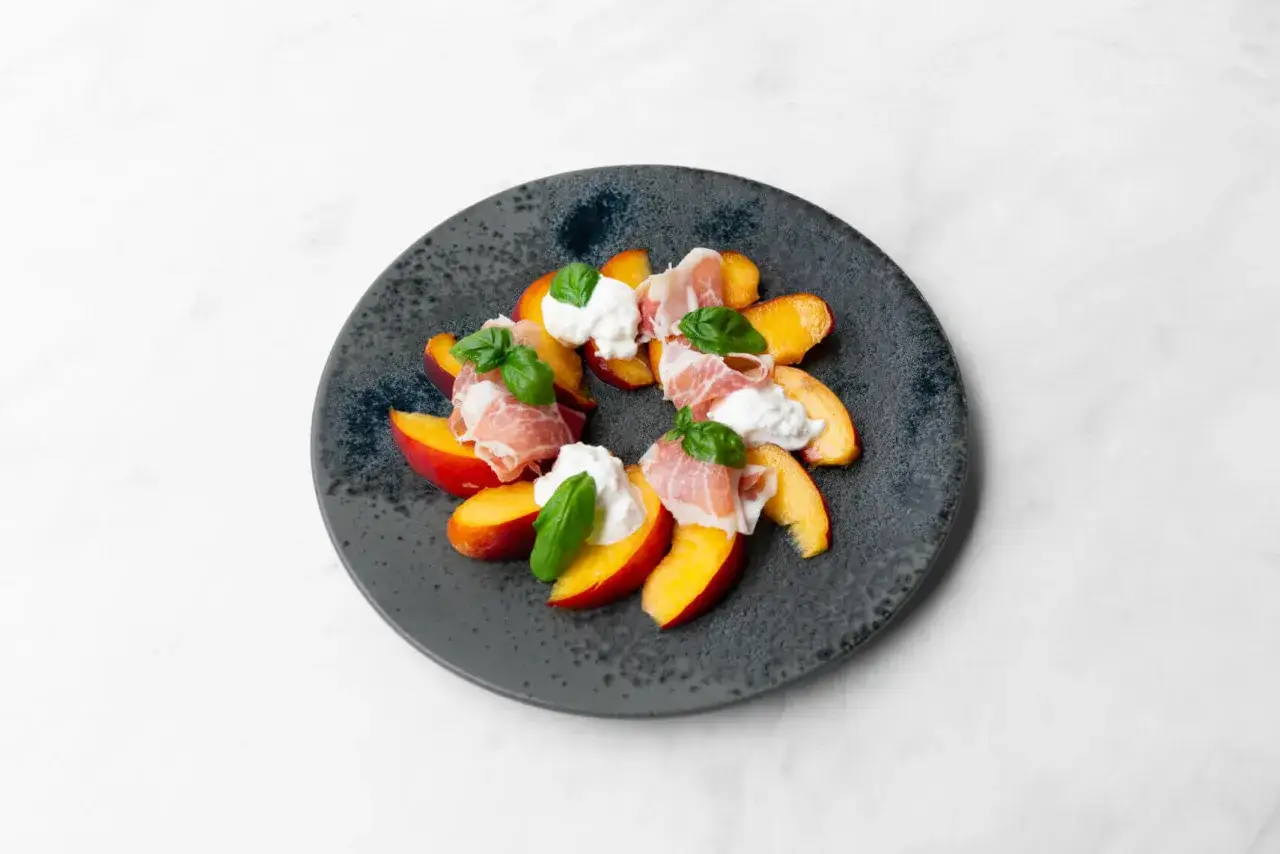 Ein Teller mit Pfirsichspalten, belegt mit Burrata, Prosciutto und Basilikum. Ein einfaches Gericht, das zeigt, wie vielseitig Burrata-Rezepte sein k&ouml;nnen.