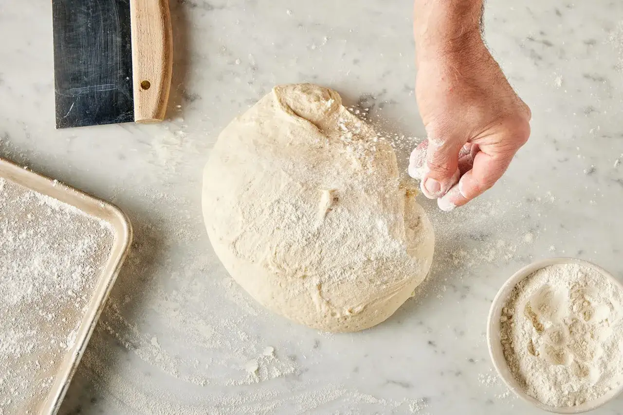 Hand streut Mehl &uuml;ber Teig. Ein Blech und eine Sch&uuml;ssel mit Mehl liegen daneben.