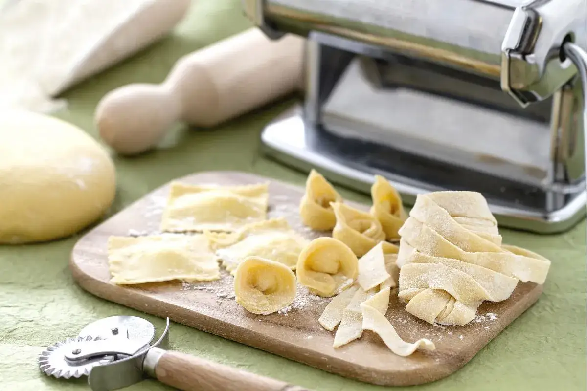Frischer Teig f&uuml;r Pasta Rezepte: Ravioli, Tortellini und Tagliatelle liegen bereit. Eine Nudelmaschine und ein Teigr&auml;dchen sind im Hintergrund zu sehen.