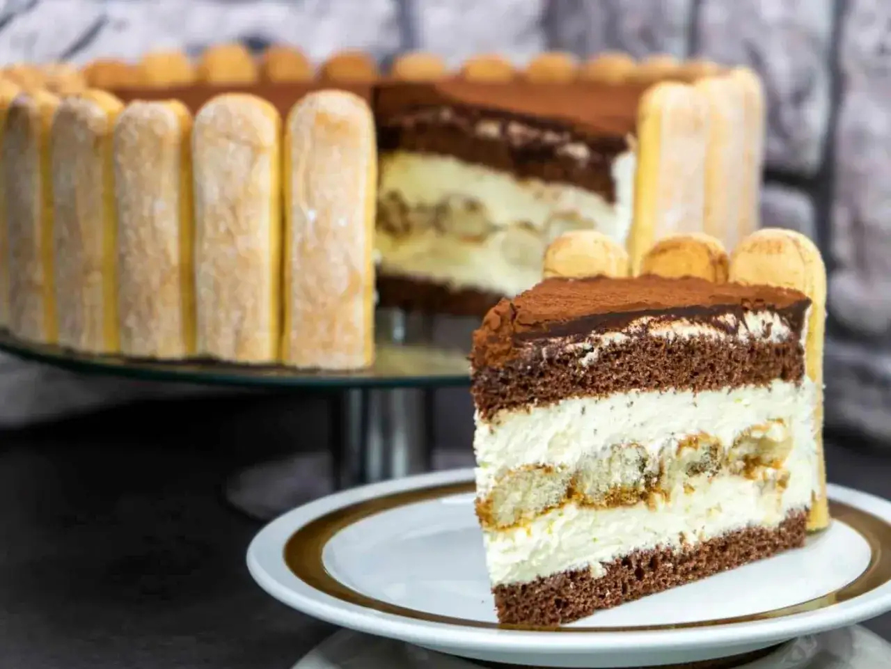 Ein Stück Tiramisu Torte mit Löffelbiskuits und Kakao bestäubt. Im Hintergrund die ganze Torte, ebenfalls mit Löffelbiskuits umrandet.