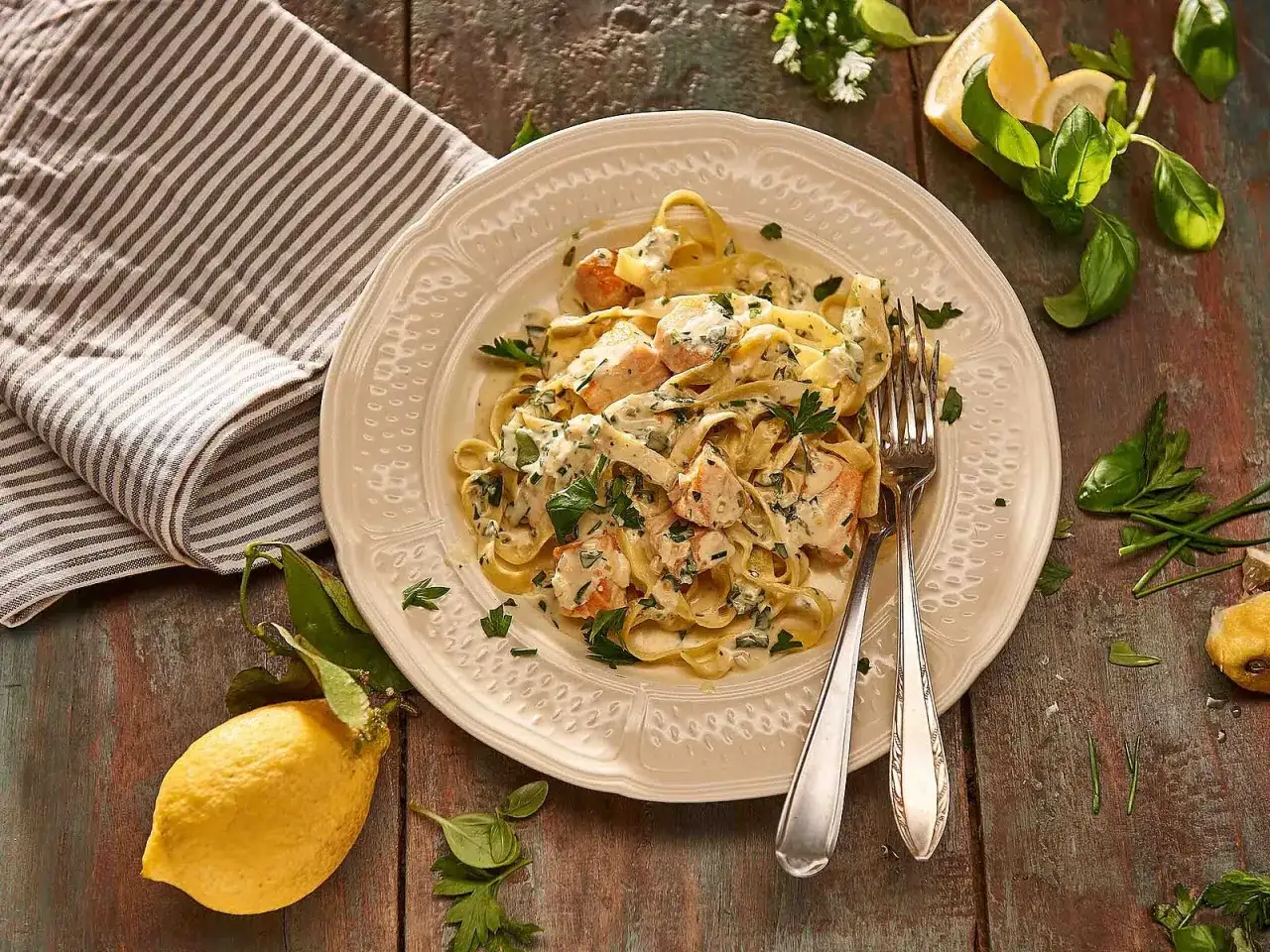 Cremige Pasta al Salmone mit Lachs, Kr&auml;utern und Zitrone auf einem Teller, daneben eine Serviette und frische Zitronenspalten.