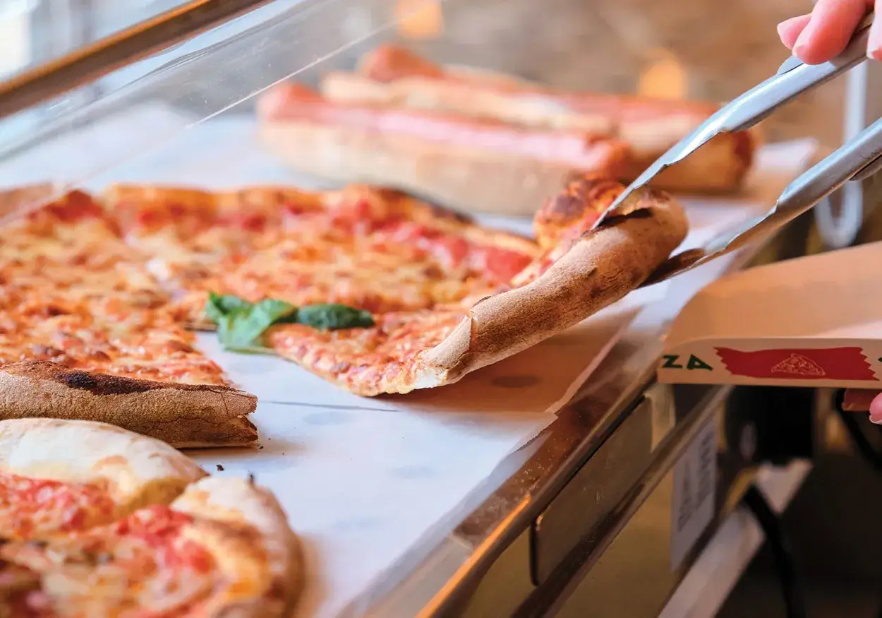 Eine Zange hebt ein Stück New York Style Pizza aus der Auslage. Der Käse schmilzt, die Kruste ist goldbraun.
