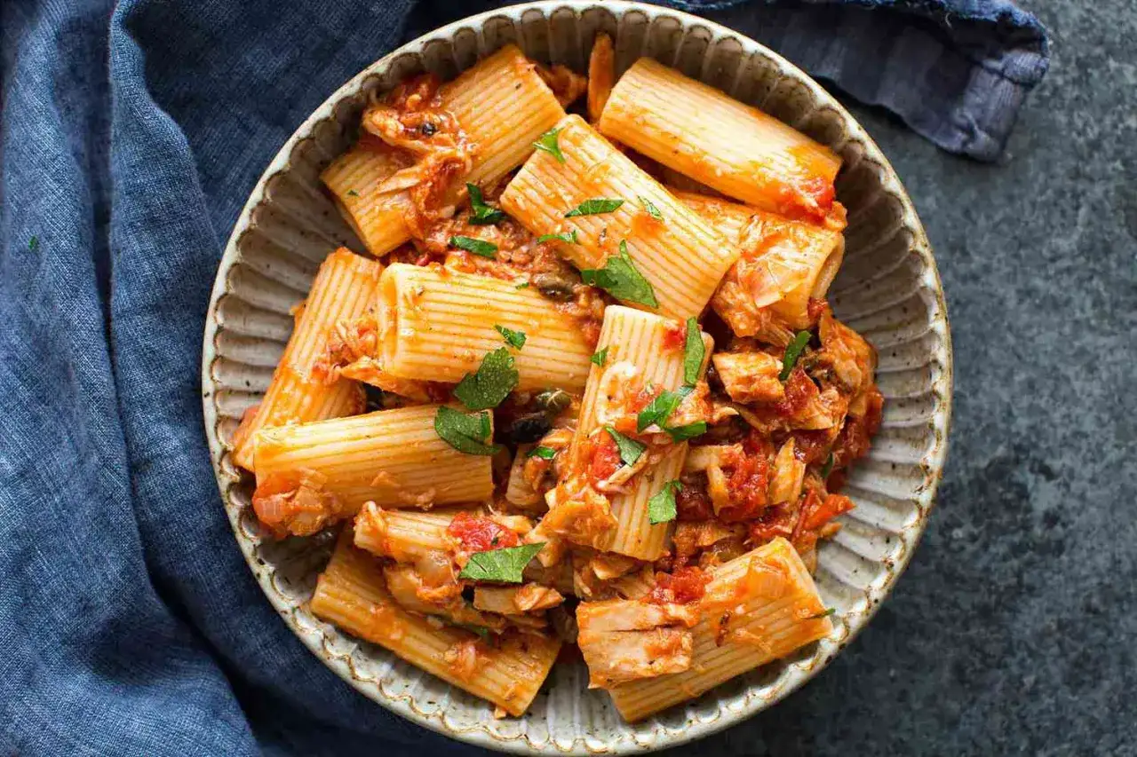 Herzhafte Thunfisch Pasta mit Tomatensauce und frischer Petersilie in einer rustikalen Sch&uuml;ssel, serviert auf einem blauen Tuch.