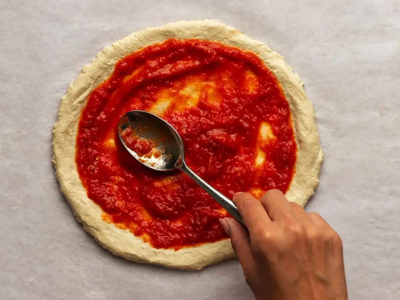 Eine Hand verteilt mit einem L&ouml;ffel Tomatensauce auf einem Pizzateig. Die Menge der Sauce pro Person ist entscheidend f&uuml;r den perfekten Geschmack.