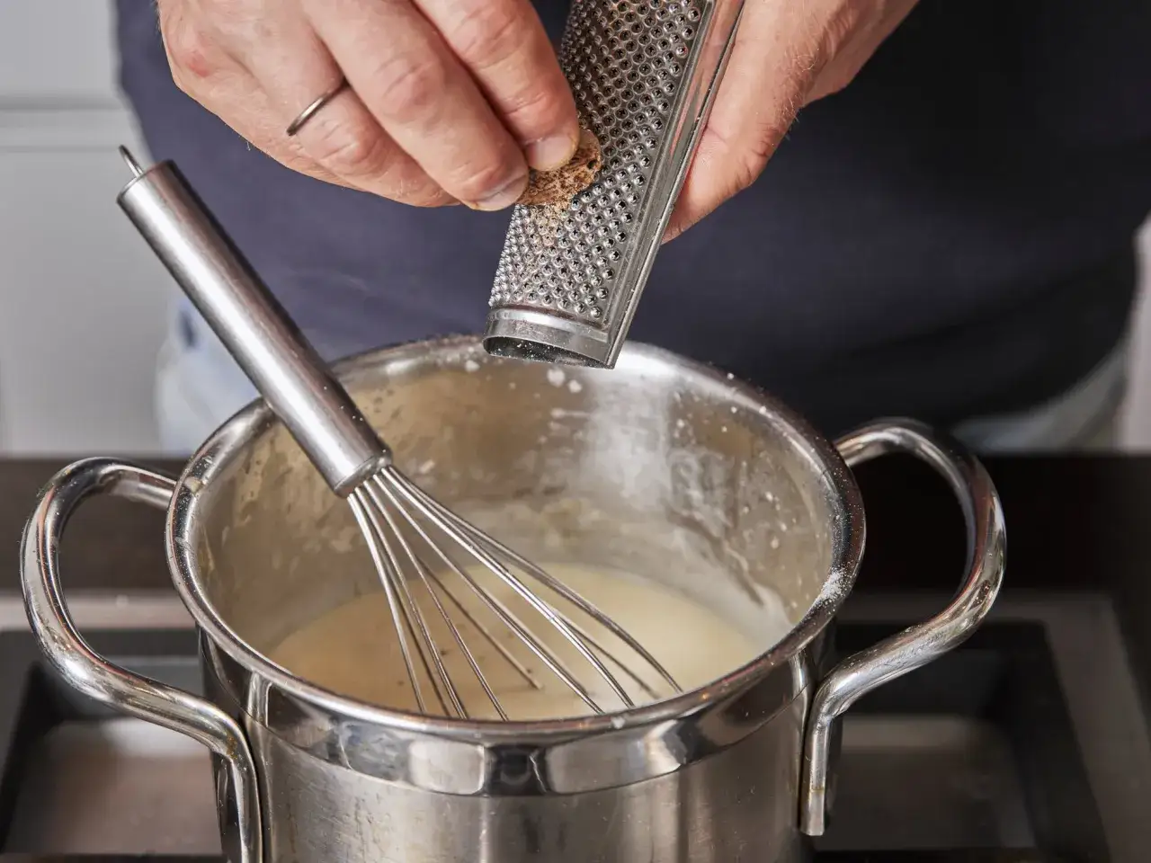 Frisch geriebene Muskatnuss verfeinert die cremige Bechamelsauce f&uuml;r die Lasagne. Ein Schneebesen r&uuml;hrt die Sauce in einem Topf.