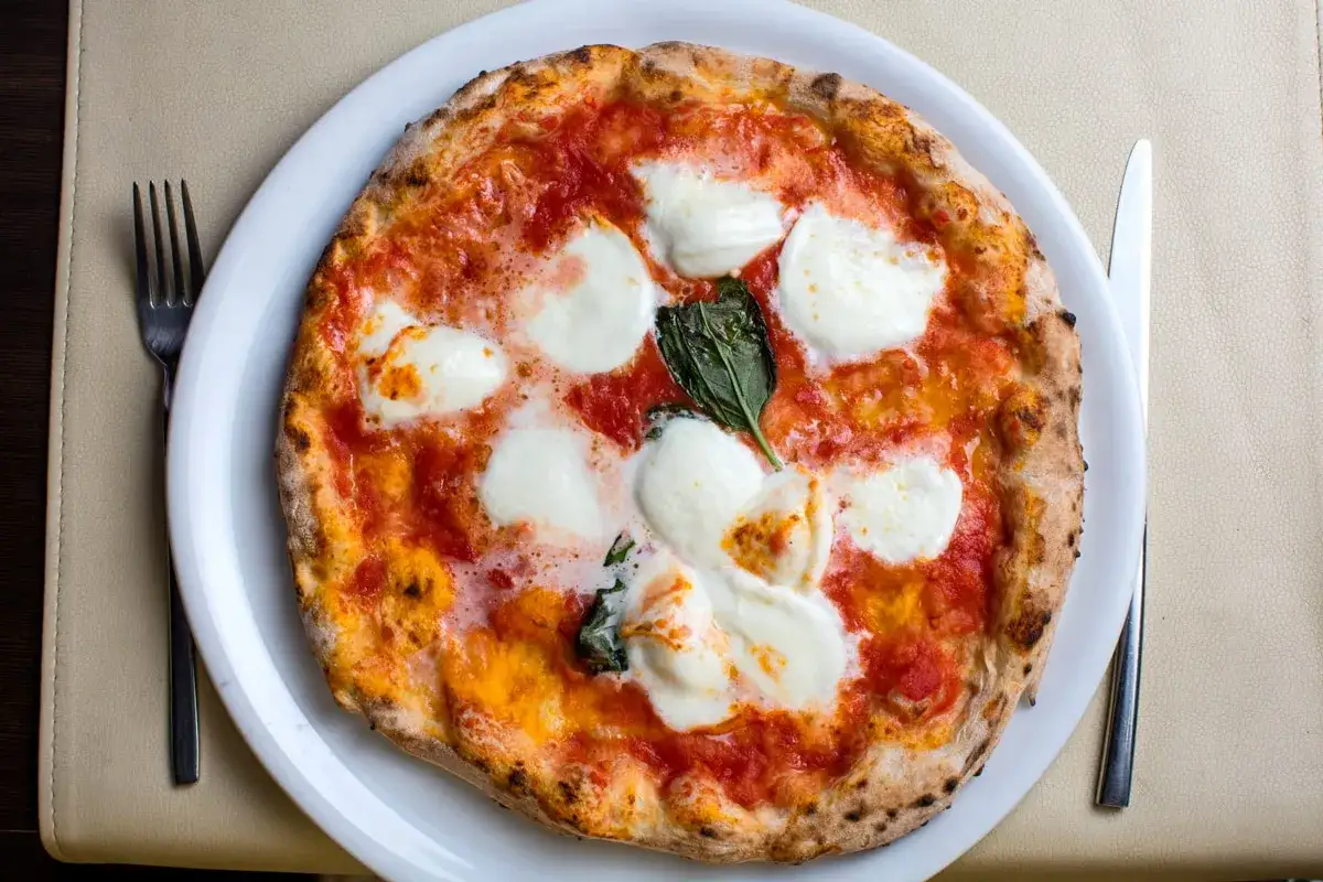 Eine Margherita-Pizza mit geschmolzenem Mozzarella, Tomatensauce und Basilikum auf einem knusprigen, italienischen Pizzateig. Gabel und Messer liegen bereit.