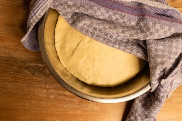 Frischer, aufgegangener Pizzateig ruht in einer Schüssel, bedeckt mit einem gemusterten Tuch. Perfekt für Ihr Pizzateig Rezept.