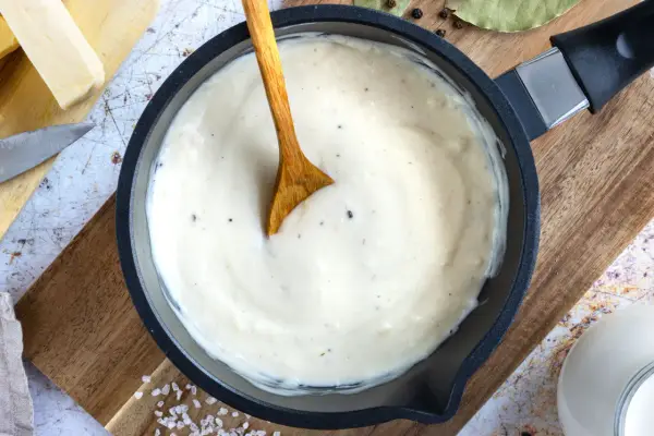 Cremige Bechamelsauce in einer Pfanne, bereit für die Lasagne. Ein Holzlöffel rührt die sämige Masse.