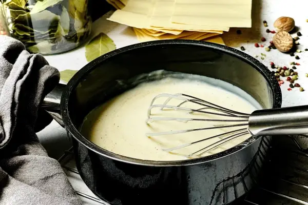 Cremige Béchamelsauce wird in einem Topf mit einem Schneebesen zubereitet. Daneben liegen Lasagneplatten und Gewürze.