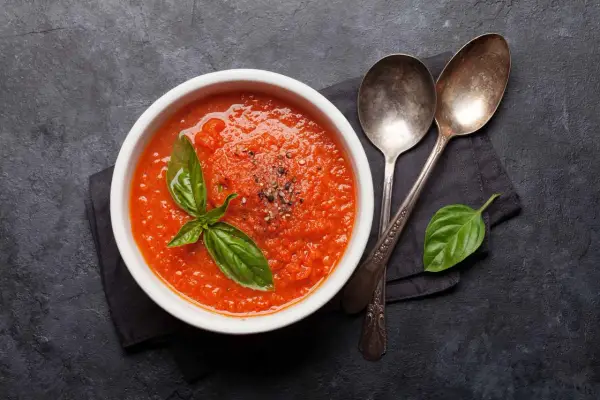 Schüssel mit selbstgemachter Tomatensauce, garniert mit Basilikum und Pfeffer, daneben zwei Löffel auf dunklem Hintergrund.