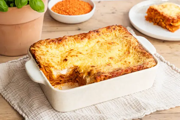 Herzhafte Linsen Lasagne mit goldbraun überbackener Käseschicht, daneben eine Schüssel roter Linsen und ein Teller mit einem Stück Lasagne.